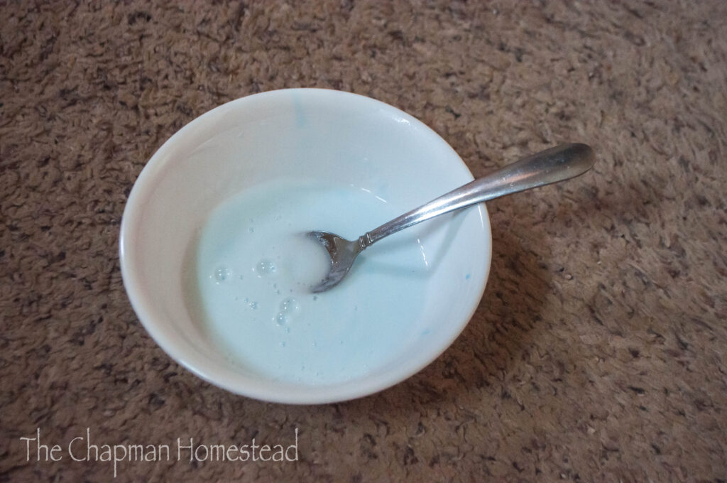 Photo of bowl with spoon inside of it. Baking soda, blue dawn, and water have been mixed together to form light blue paste.