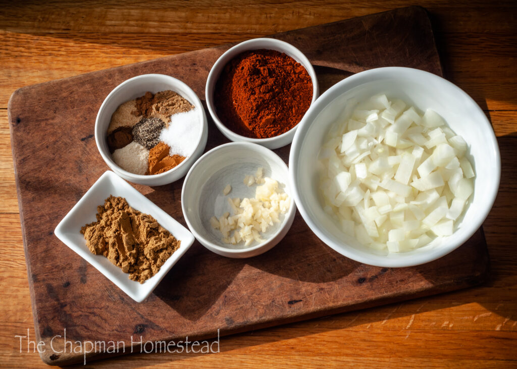 Photo with many different white bowls. One bowl contains chili powder, one contains cumin, one contains all the other dried spices used, one contains minced garlic, and one contains diced onion.